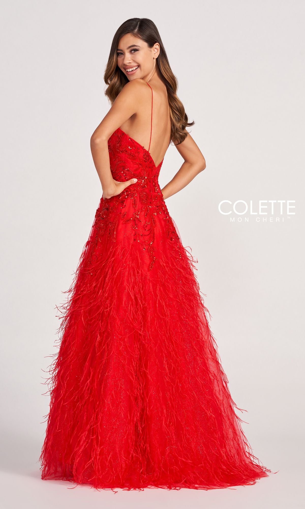 A woman poses, smiling over her shoulder in the Colette CL2044 Feather Prom Ball Gown—a long red v-neck dress with a low back, intricate feather detailing, and a flared skirt.